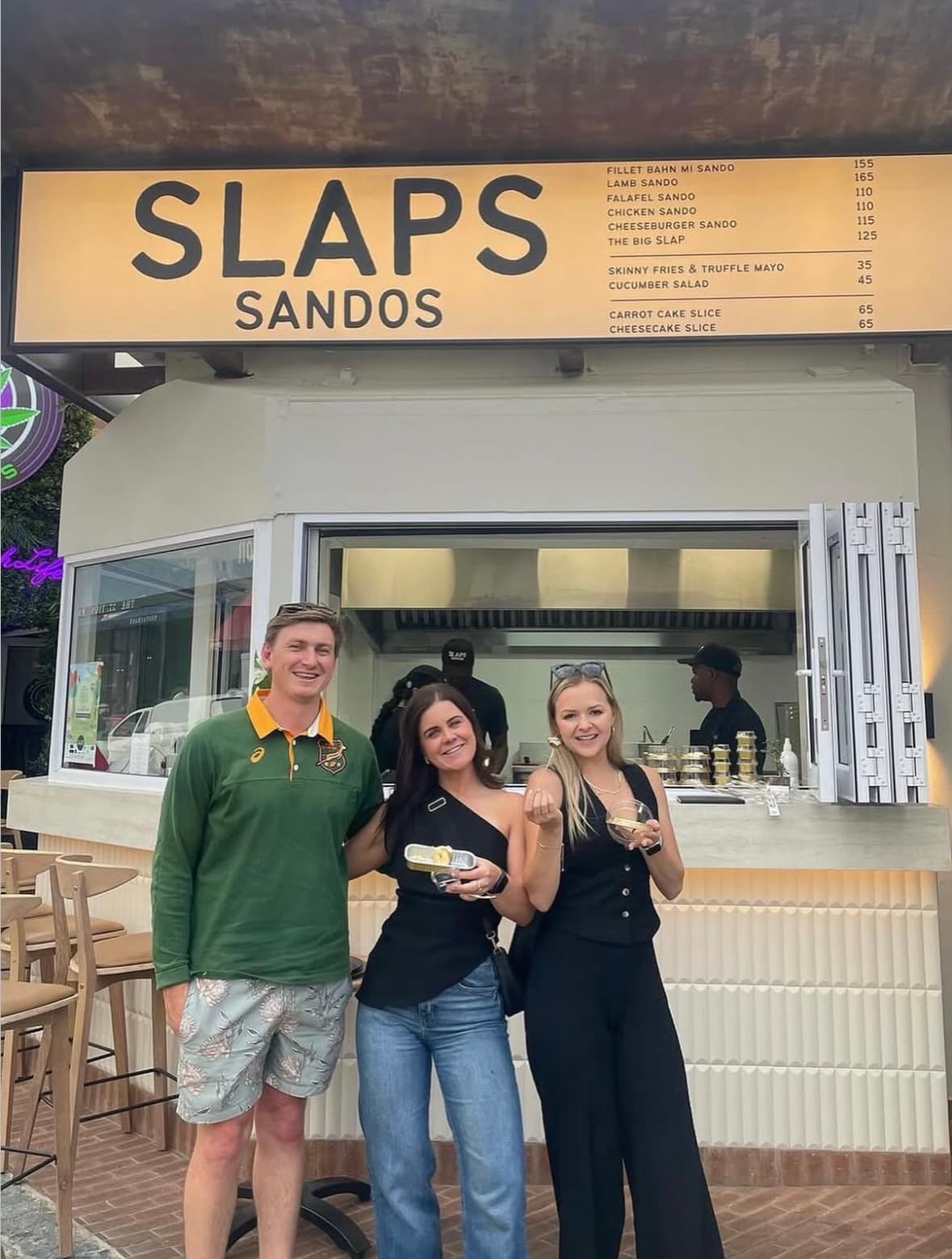 Three friends hanging out in front of the SLAPS Sandos sign with their food