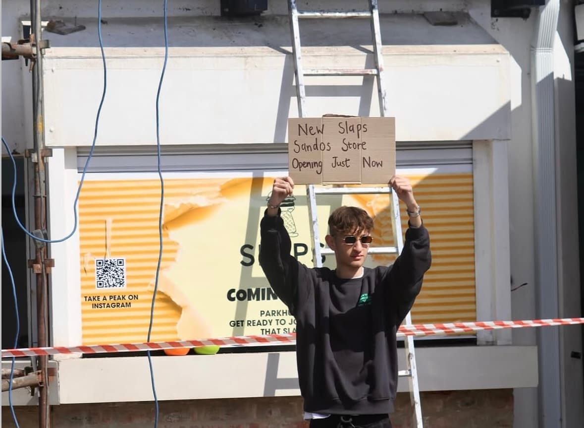 A passerby holding a cardboard sign announcing the new SLAPS Sandos store opening