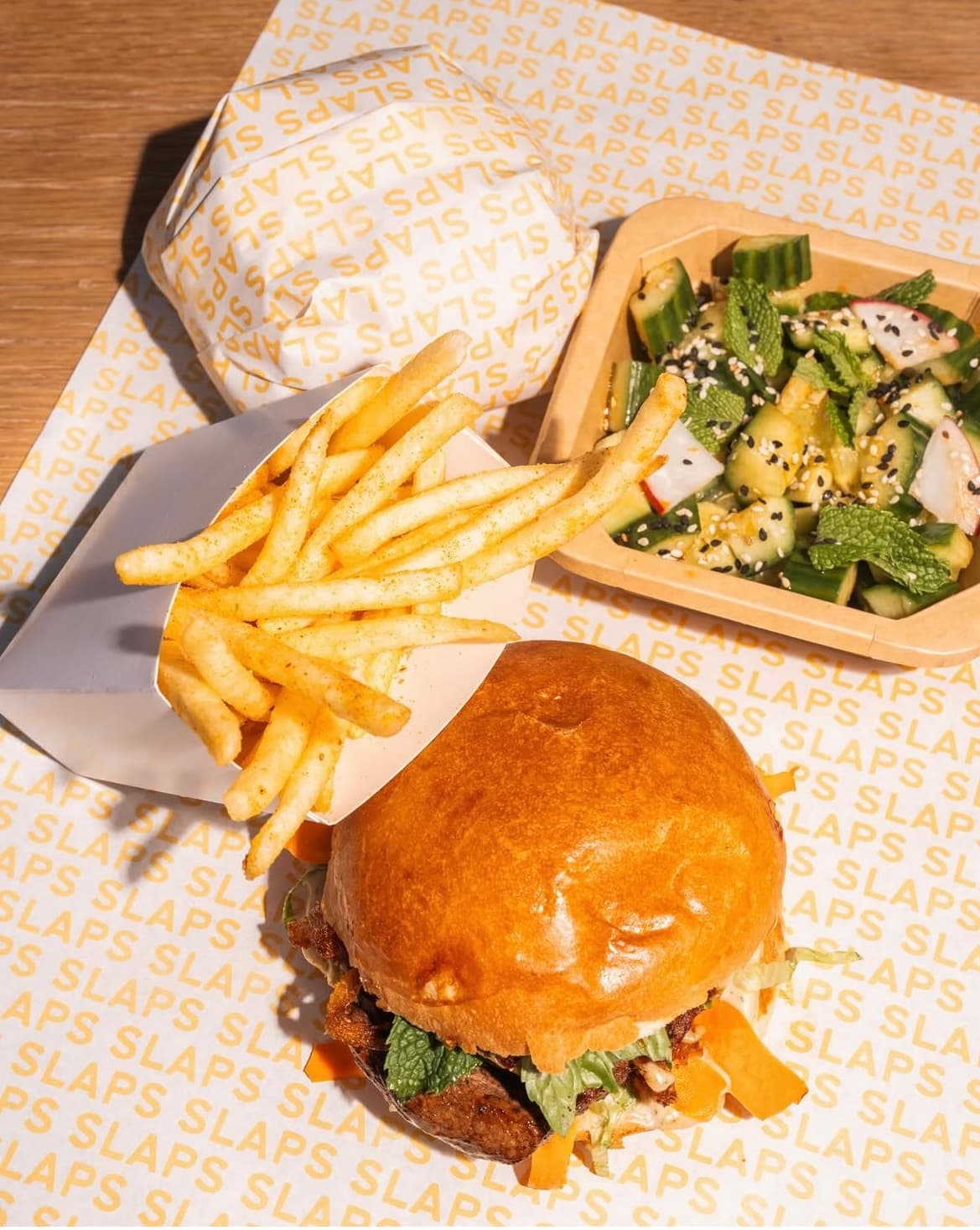 A SLAPS sando with skinny fries and cucumber salad on the yellow SLAPS wrapping paper