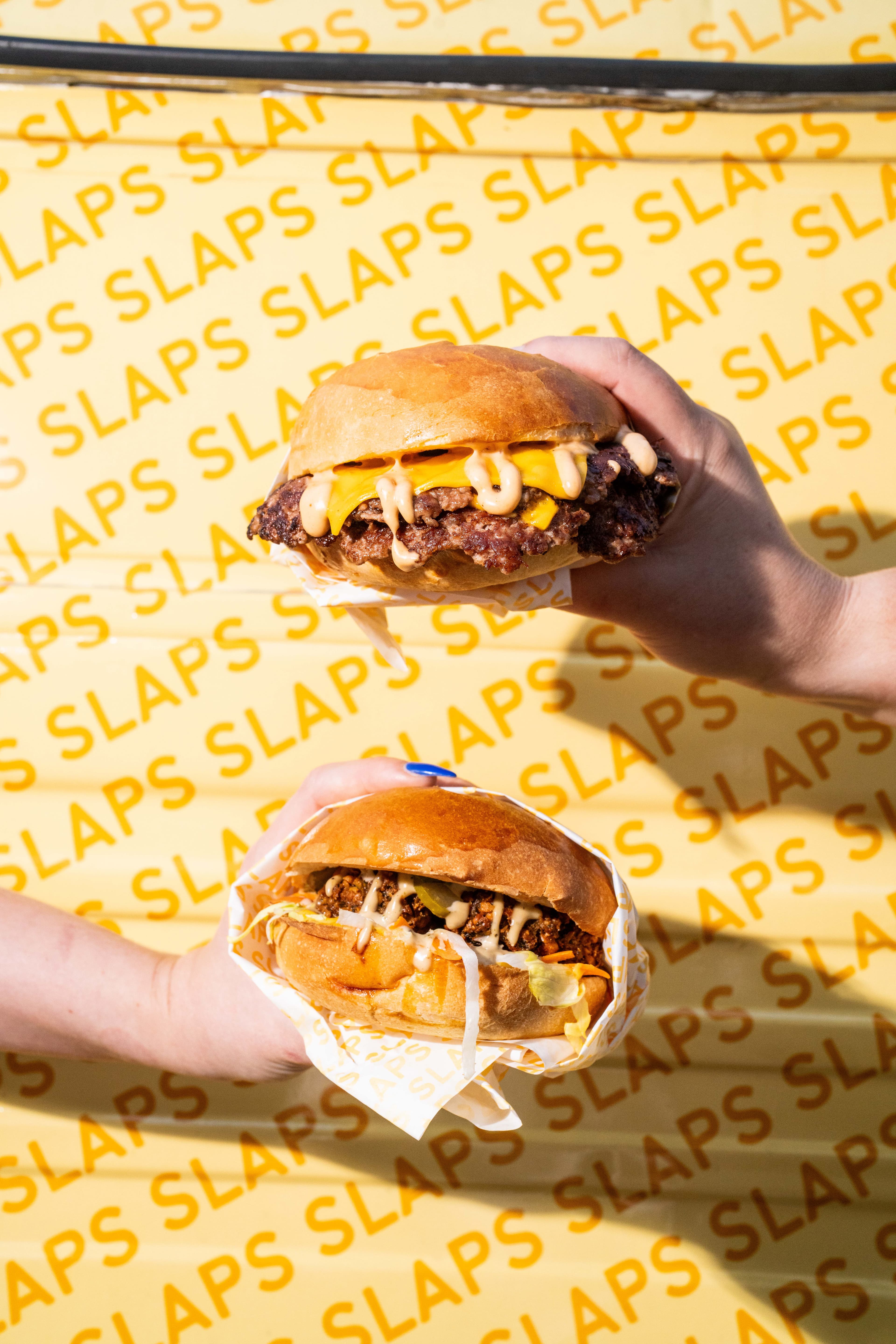 Two hands holding SLAPS burgers up against the yellow SLAPS wrapping paper